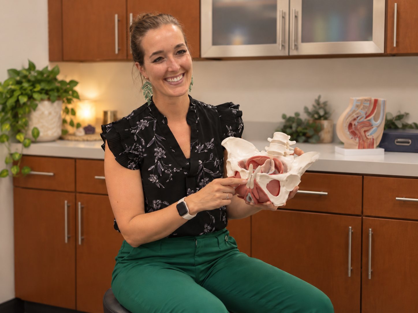 Dr. Bri seated, holding an anatomical model of the pelvis and pointing at the pelvic floor.